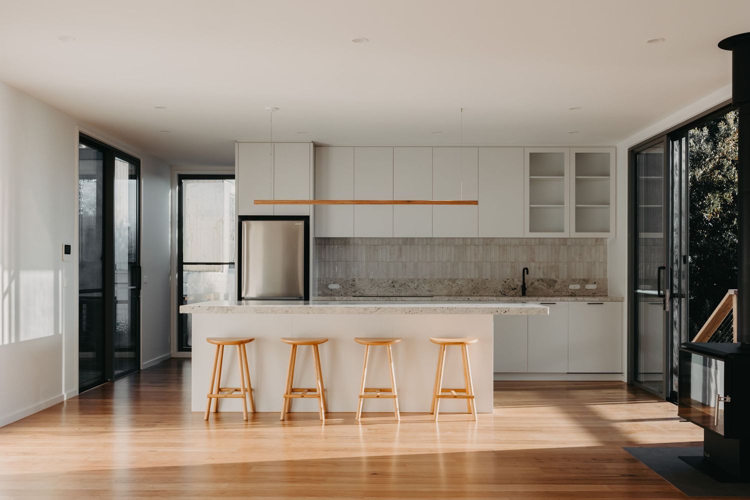 Modern white kitchen with timber bar stools, hardwood floors, glass cabinets, and stainless steel appliances in an open-plan layout.