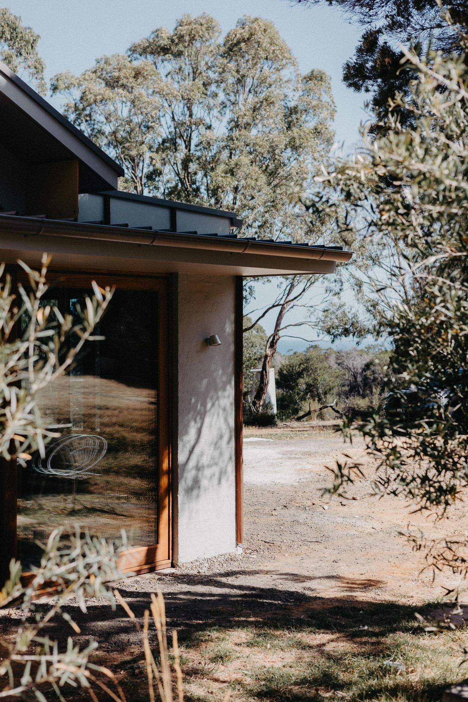 Contemporary rural home exterior with rendered walls, timber door frame and native garden under eucalyptus trees in dry landscape.