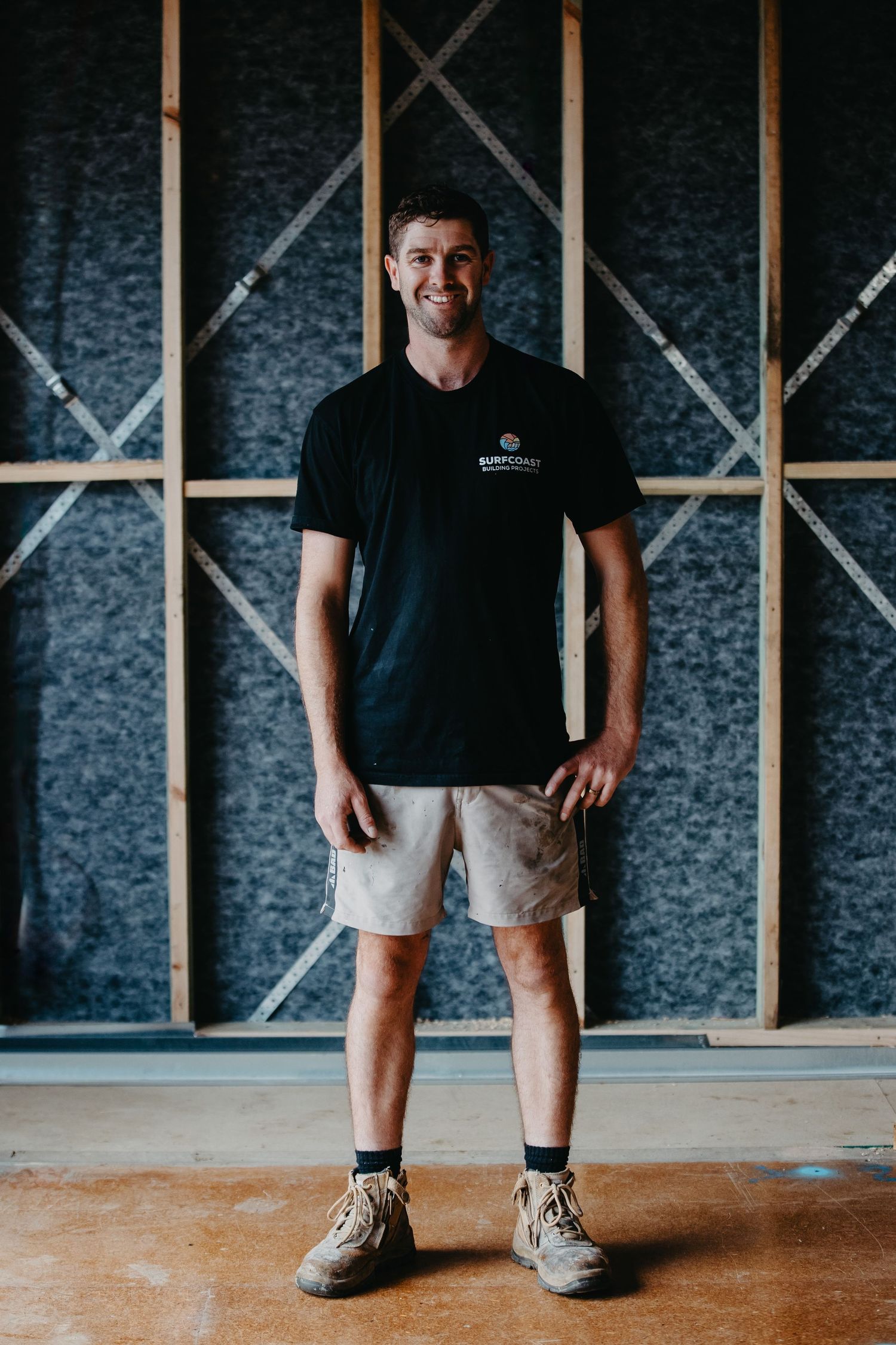 Smiling man in black t-shirt and beige shorts standing in a timber-framed room.