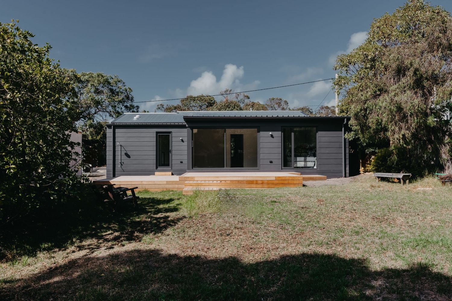 Modern dark grey single-storey home with timber deck surrounded by native Australian bush and garden landscape.