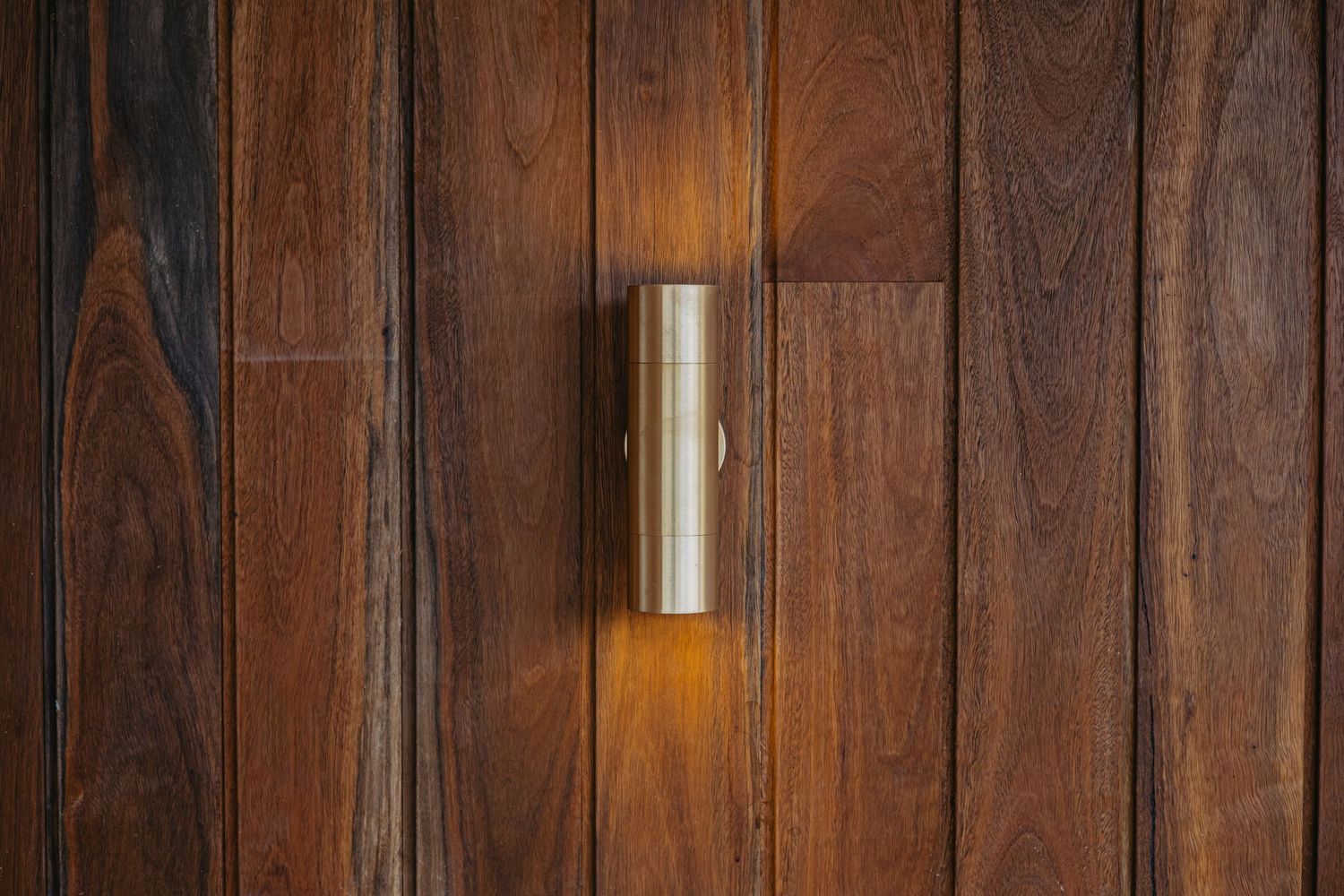 Close-up of a cylindrical brass wall light glowing warmly against rich dark timber horizontal wood cladding.