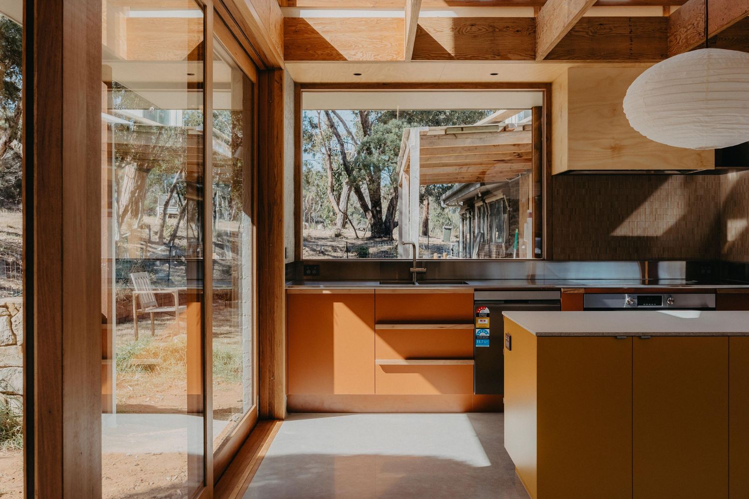 Modern kitchen with mustard yellow cabinetry, large sliding glass doors opening to timber deck with native bush surroundings.
