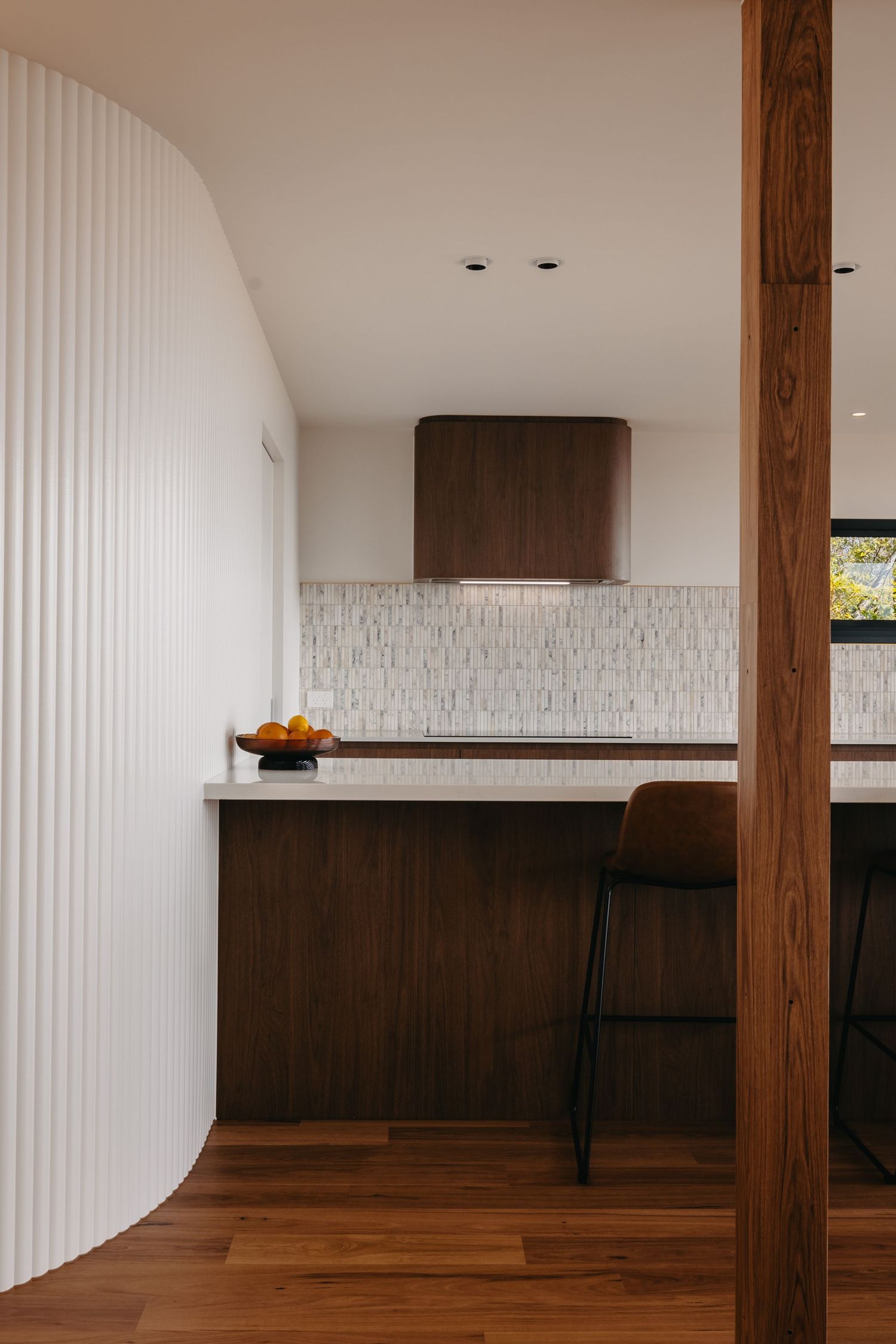 Contemporary kitchen view through curved white wall with walnut range hood and marble tile backsplash.