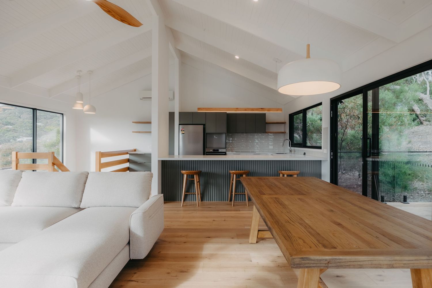 Open-plan living and dining area with vaulted ceiling, timber table, white sofa, and kitchen in background.