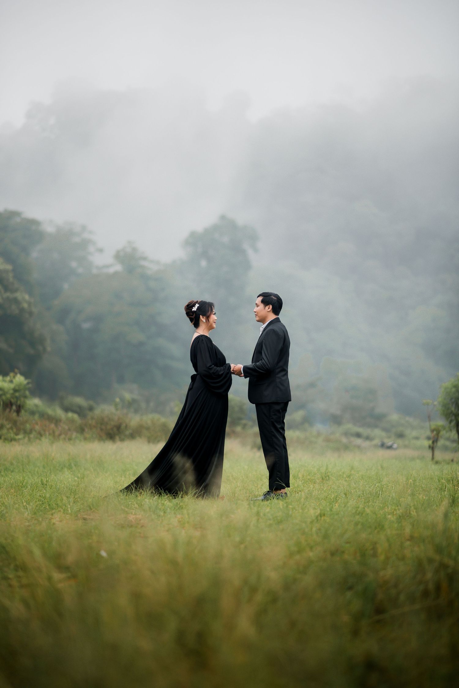 Couple in black attire holding hands in a misty green meadow with foggy forest backdrop.