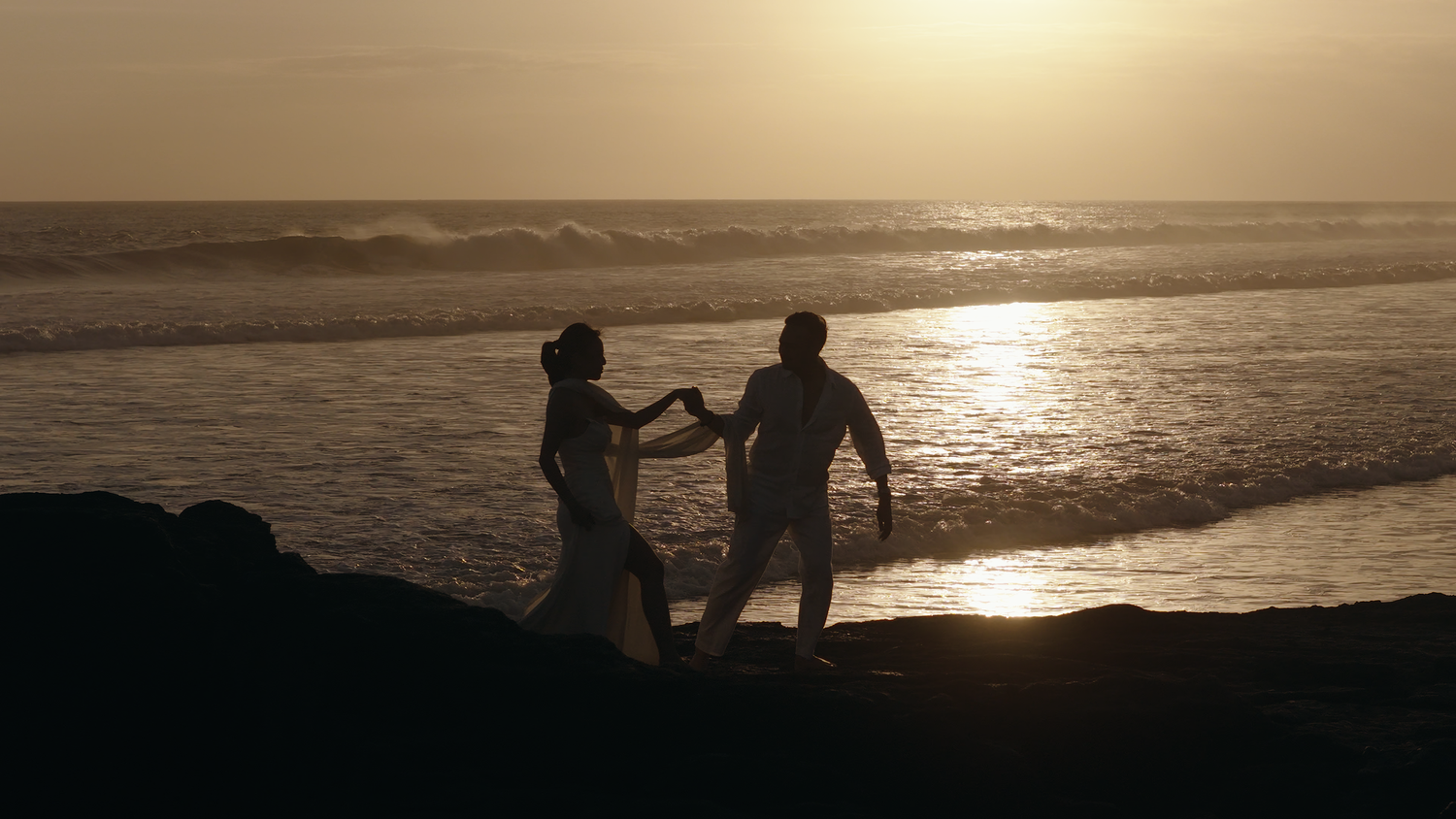 Silhouette of a couple holding hands on a rocky beach at golden sunset with ocean waves in the background.