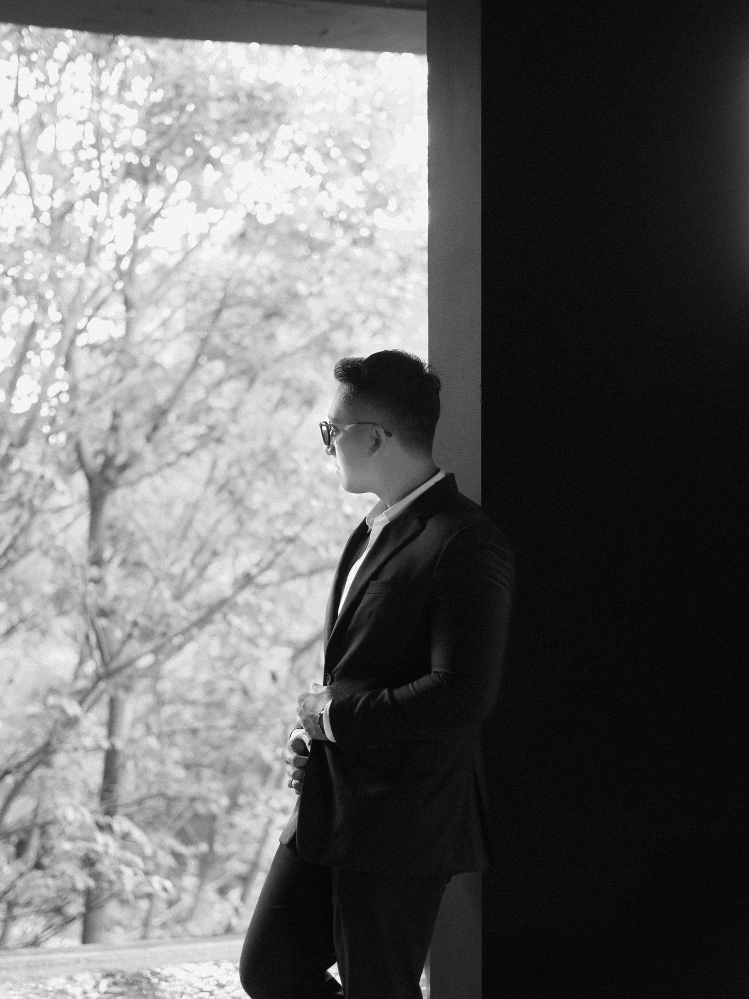 A man in a dark suit and glasses stands in profile gazing out a large window surrounded by trees in black and white.