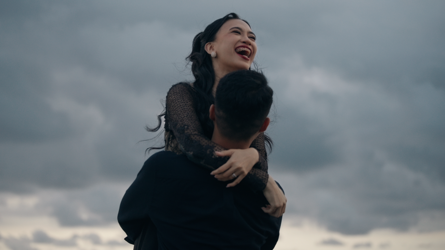 A joyful woman laughs as a man lifts her against a dramatic cloudy sky backdrop.