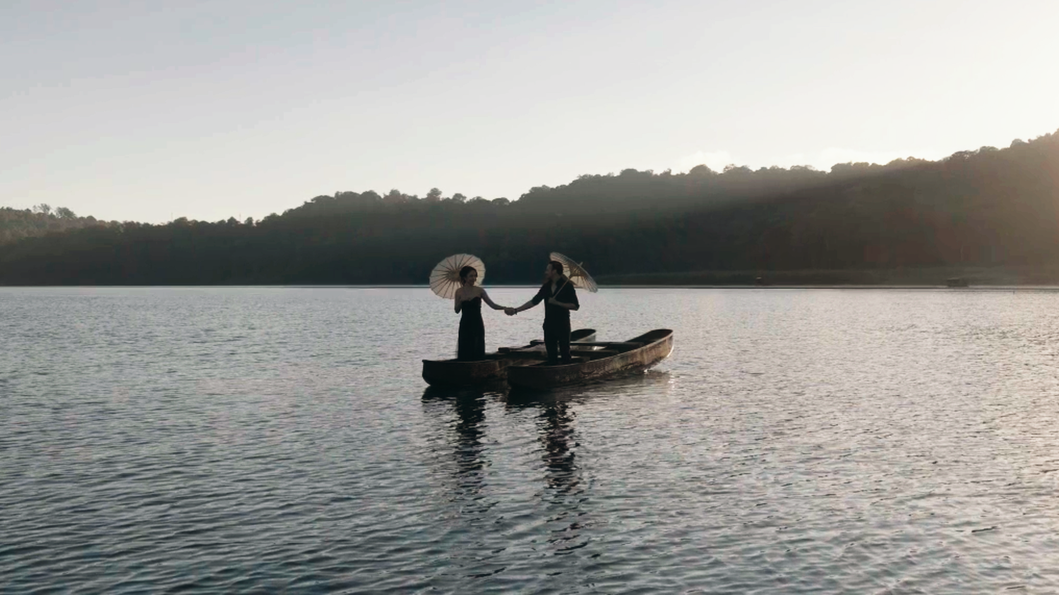 Two people in elegant attire holding hands on a canoe at sunset on a calm lake with forested hills.