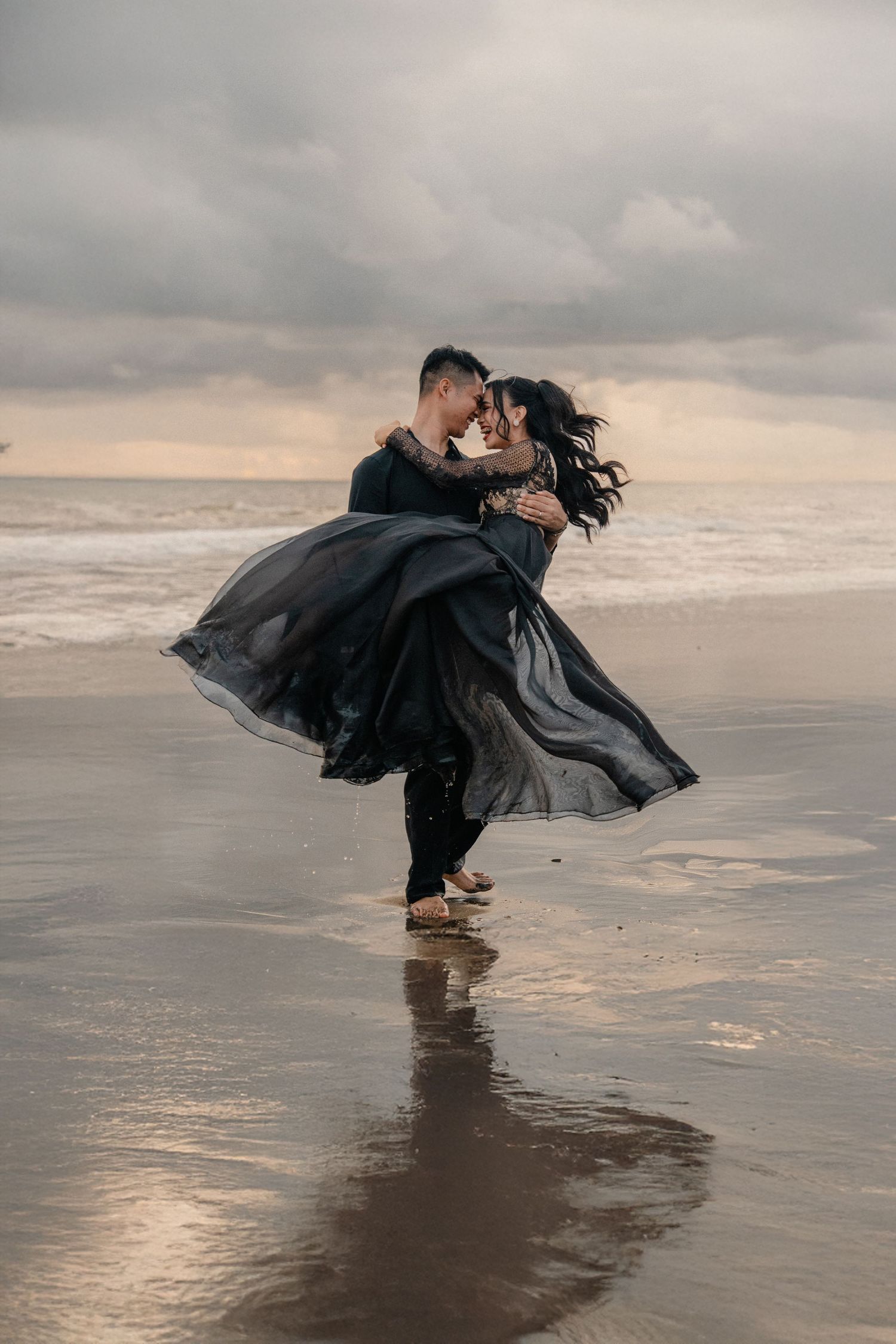 Couple in black outfits embracing on a wet beach at sunset, woman's flowing dress billowing in the wind.