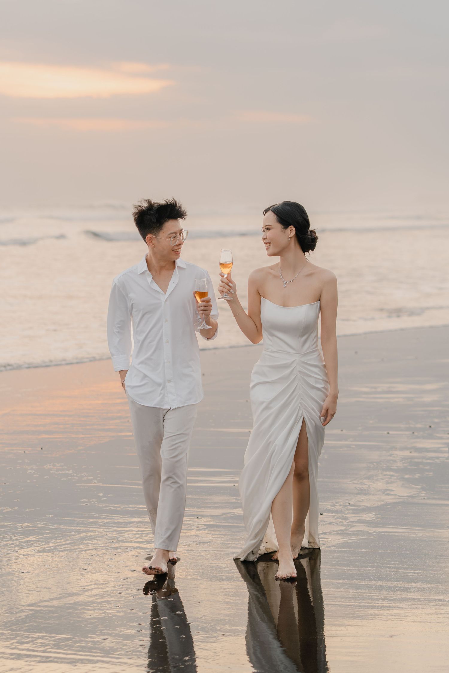 Couple in white attire toasting champagne glasses while walking on a beach at sunset.