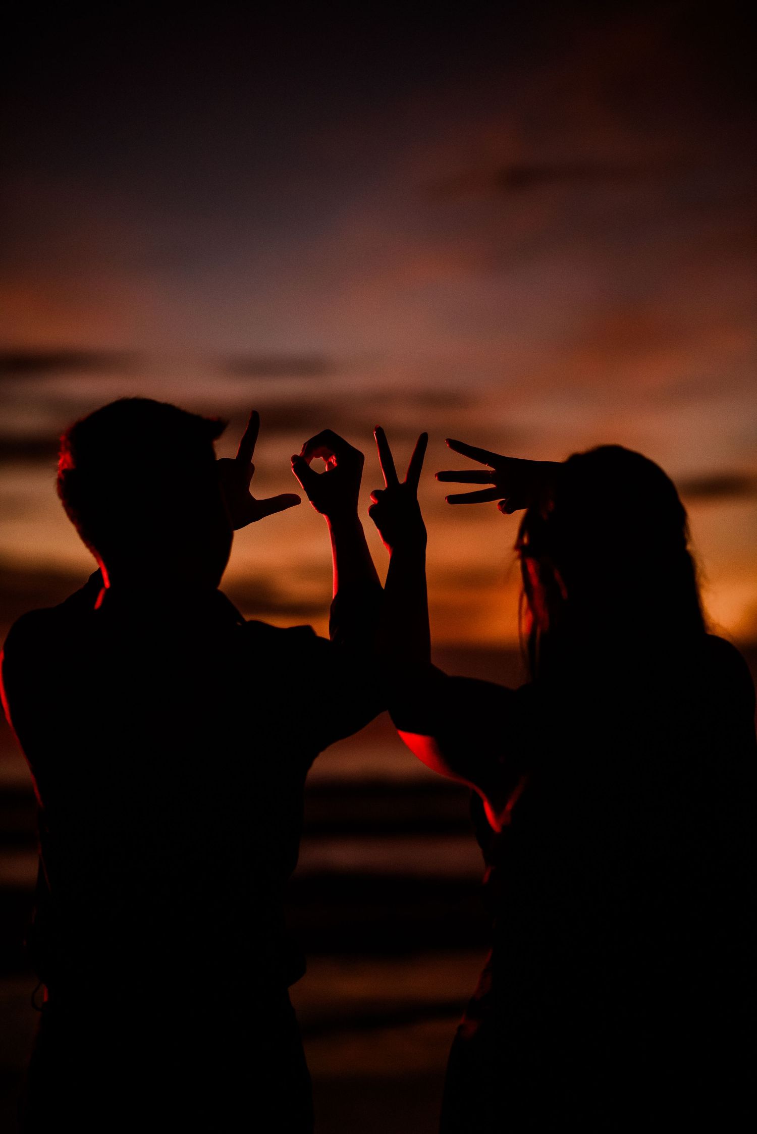 Two silhouettes making hand gestures against a vibrant orange and red sunset sky.