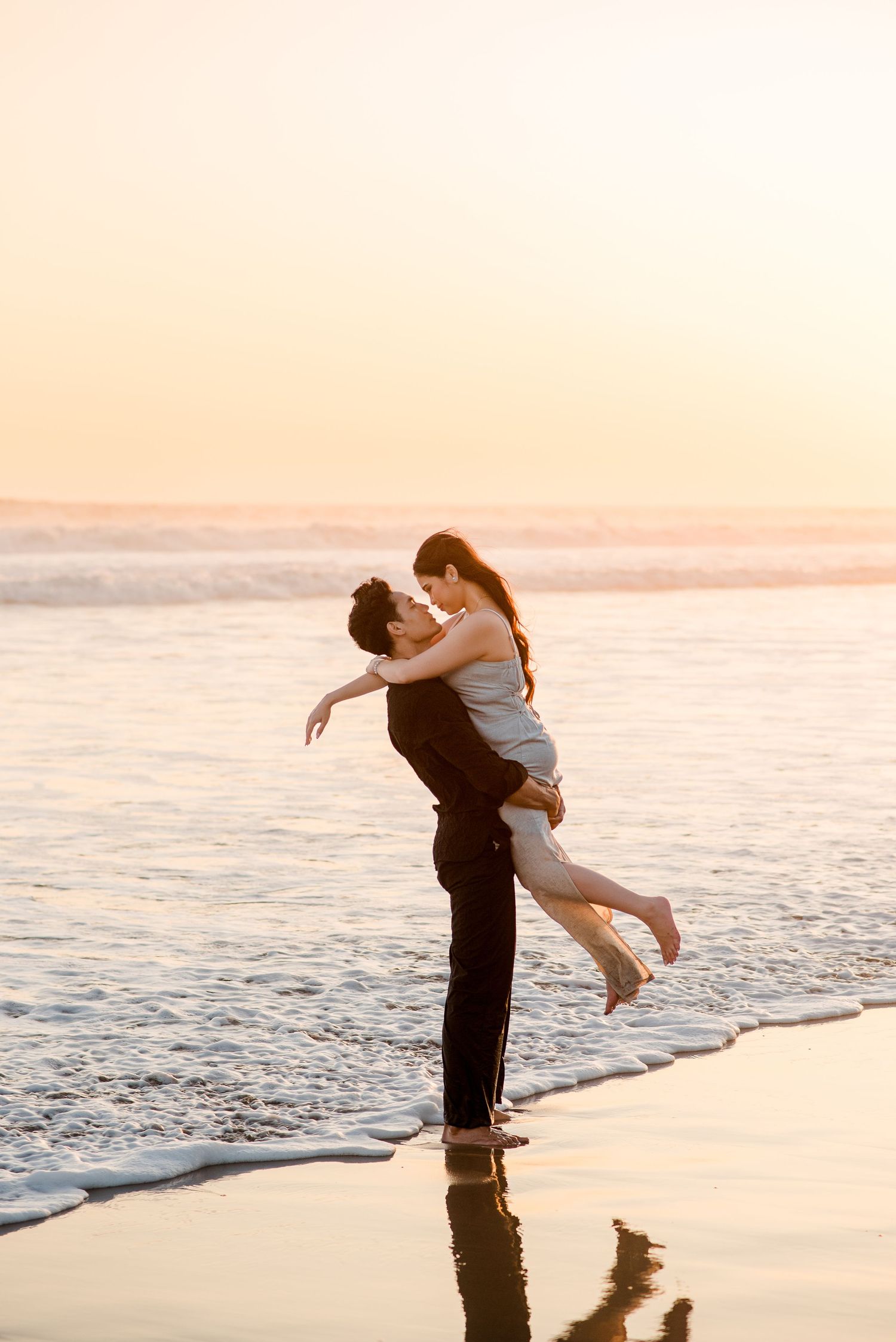 A couple shares a romantic moment at sunset on the beach, as the man lifts the woman in his arms by the ocean waves.