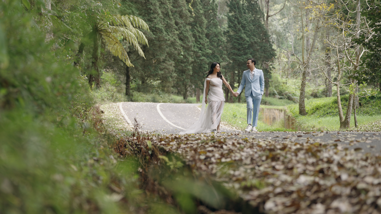 Couple holding hands walking along a scenic tree-lined path surrounded by lush green forest.