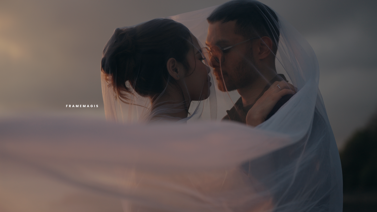 A couple shares a romantic kiss under a flowing white veil at dusk, with dramatic cloudy skies in the background.