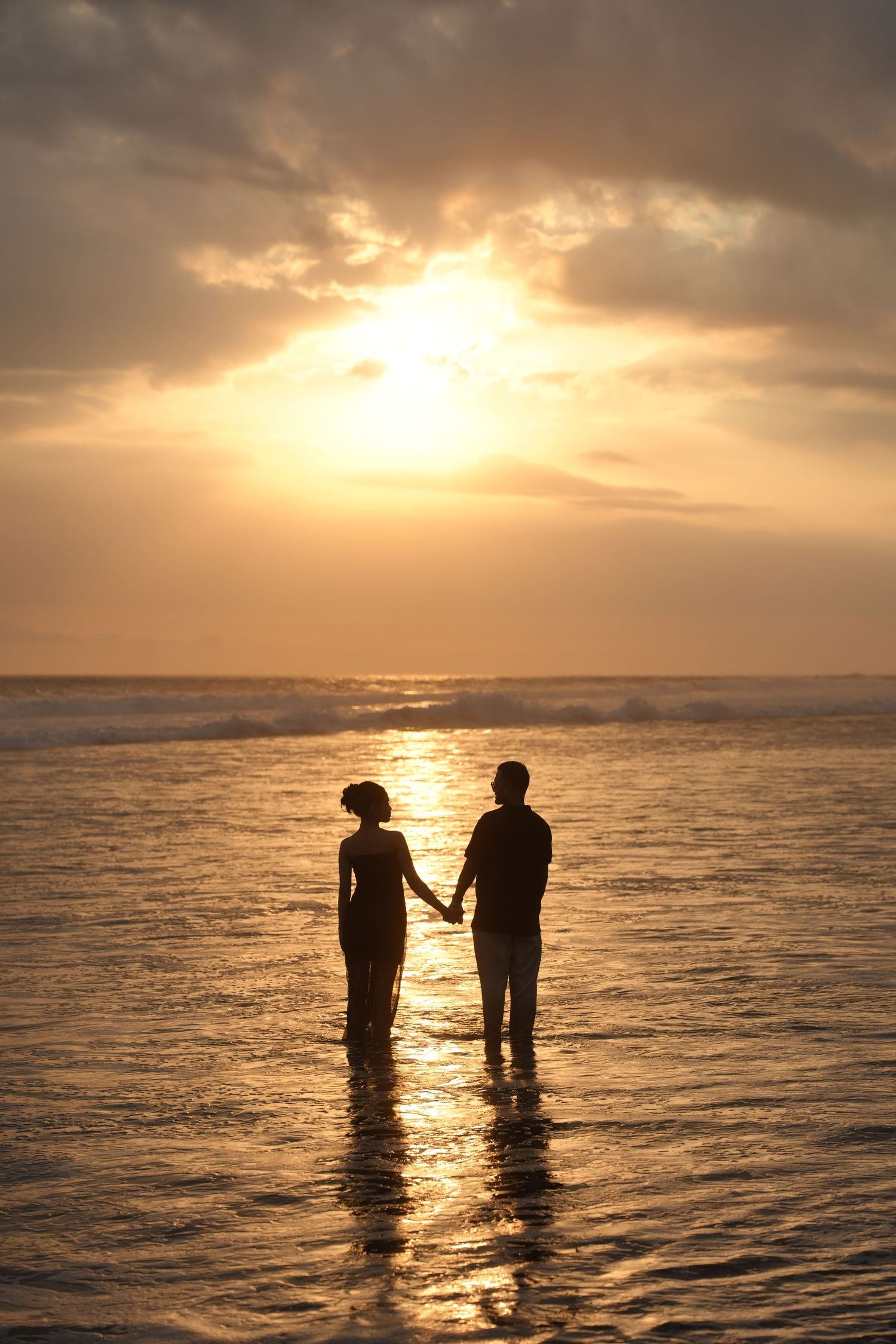 Romantic couple silhouette holding hands in ocean waves at golden sunset on tropical beach.