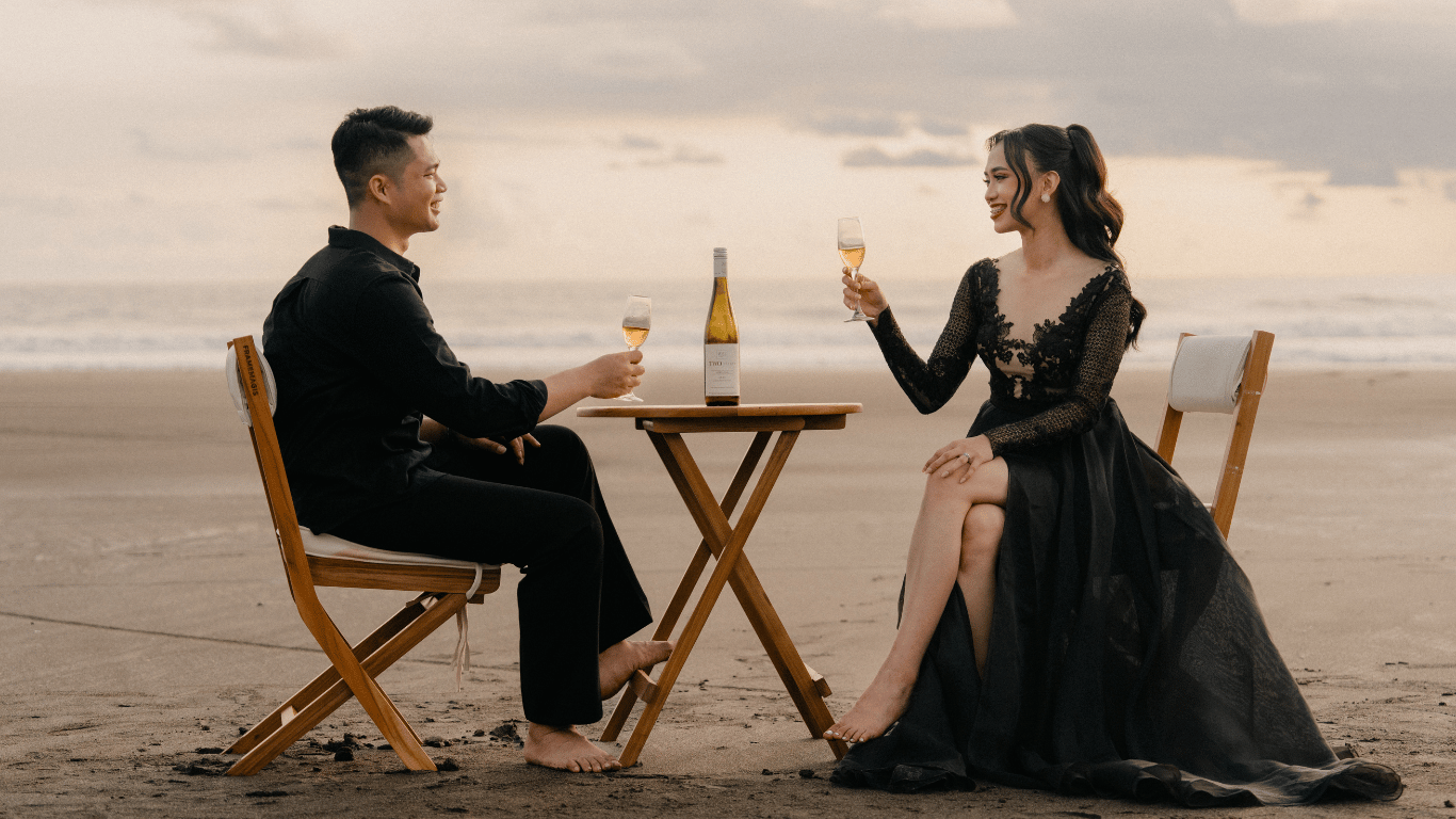 Couple in black attire sharing champagne at a romantic beach dinner table at sunset.
