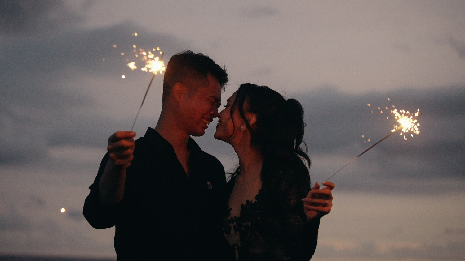 Romantic couple holding sparklers at dusk, leaning in for a kiss against a dramatic twilight sky.