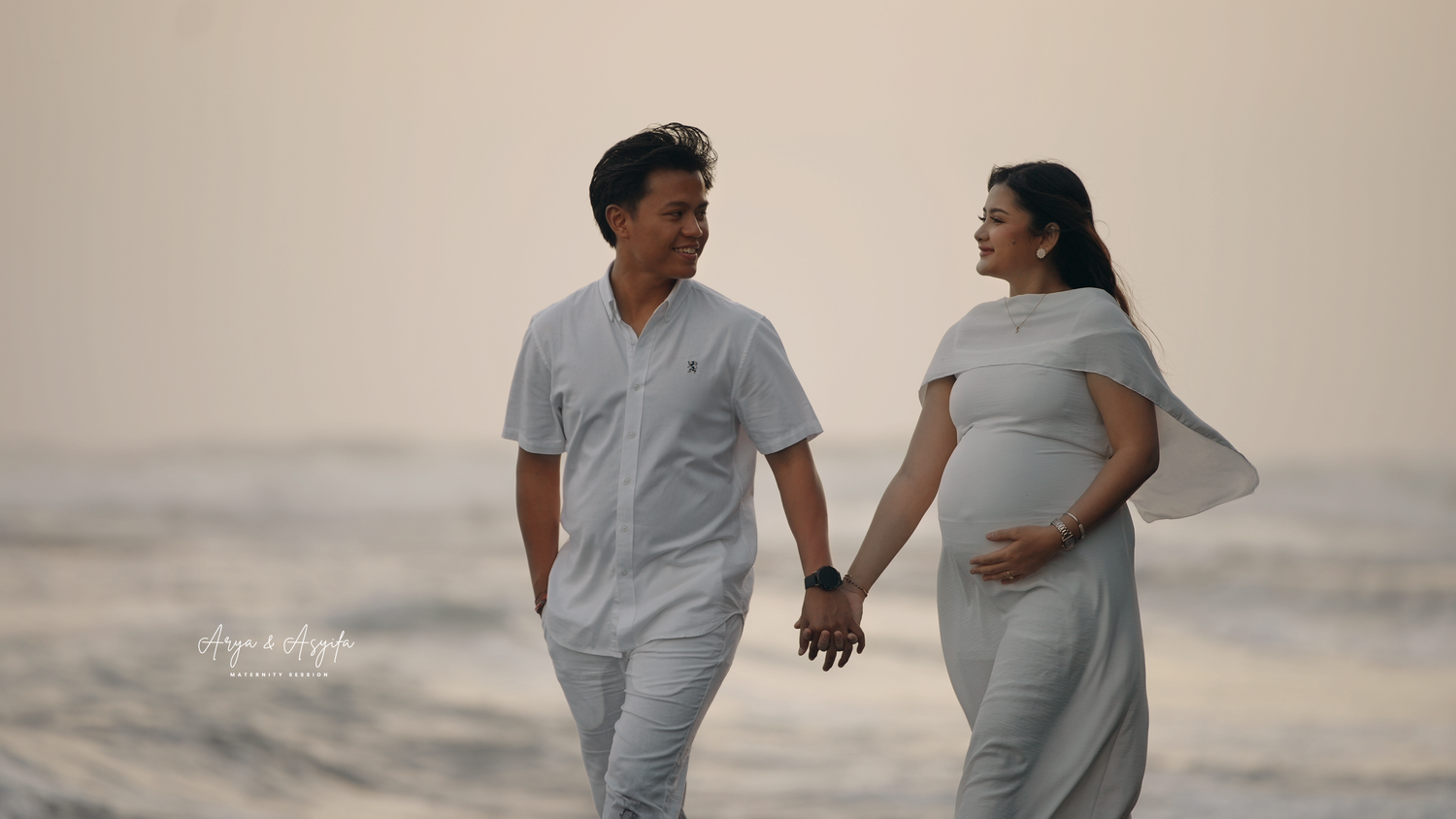 A couple holds hands on a beach at sunset, the woman in a gray maternity dress cradling her baby bump.