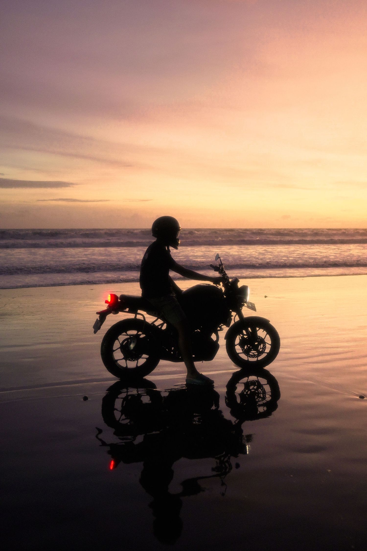 Silhouette of a motorcyclist on a wet beach at sunset, with vibrant purple and orange skies reflecting on the shore.