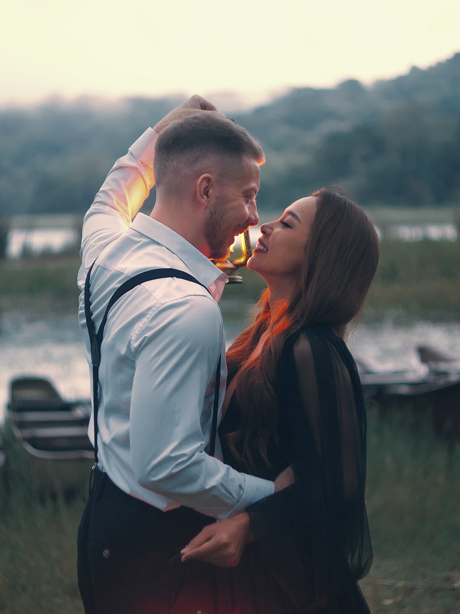 Romantic couple sharing an intimate moment near a lake at sunset, man in suspenders embracing woman in black outfit.