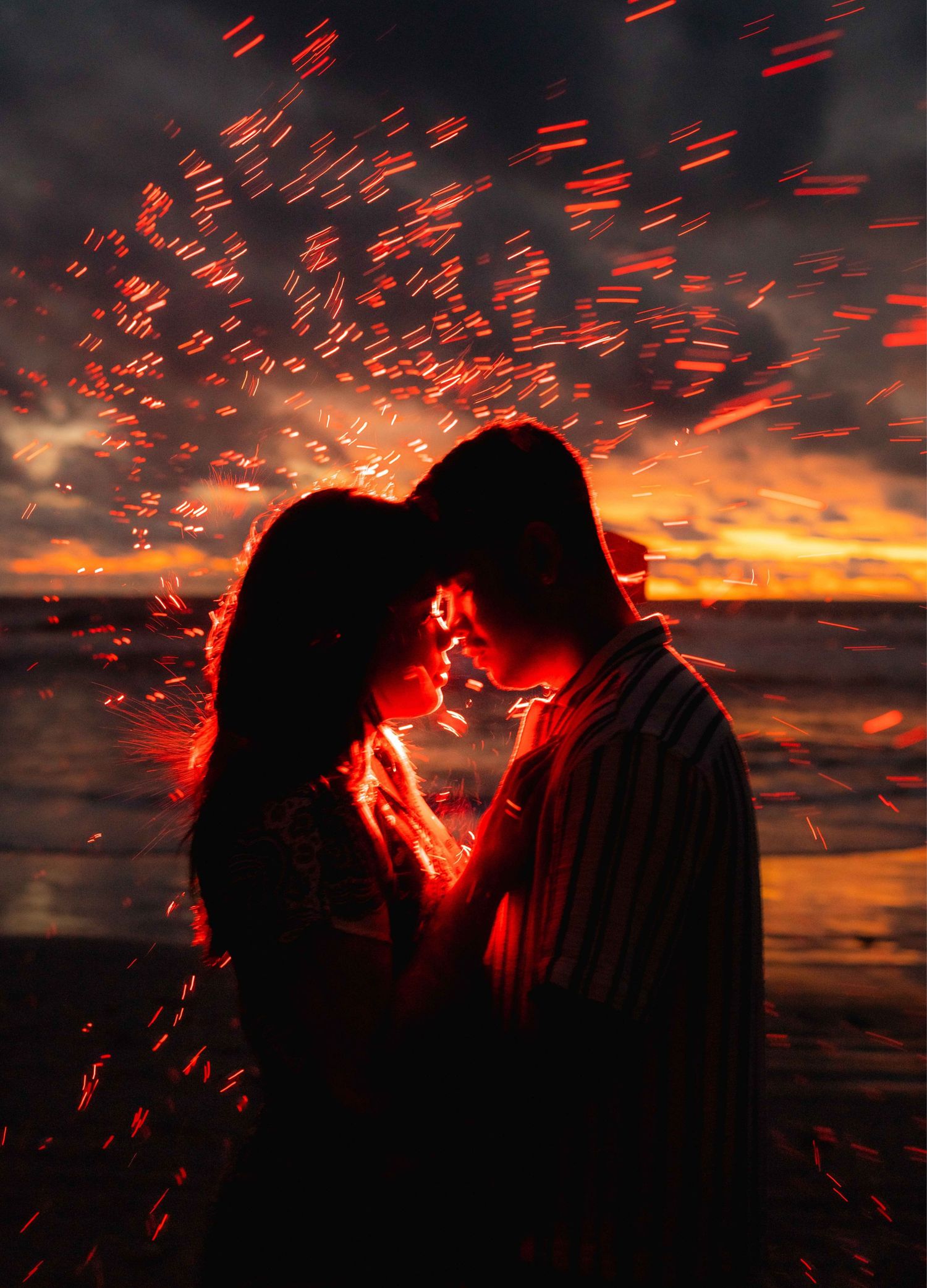 Couple sharing a romantic moment surrounded by glowing fire sparks at a dramatic sunset beach.