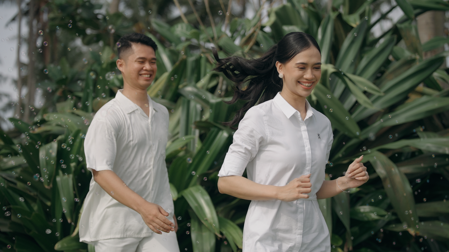 A man and woman in matching white outfits jogging together outdoors among lush green tropical plants.