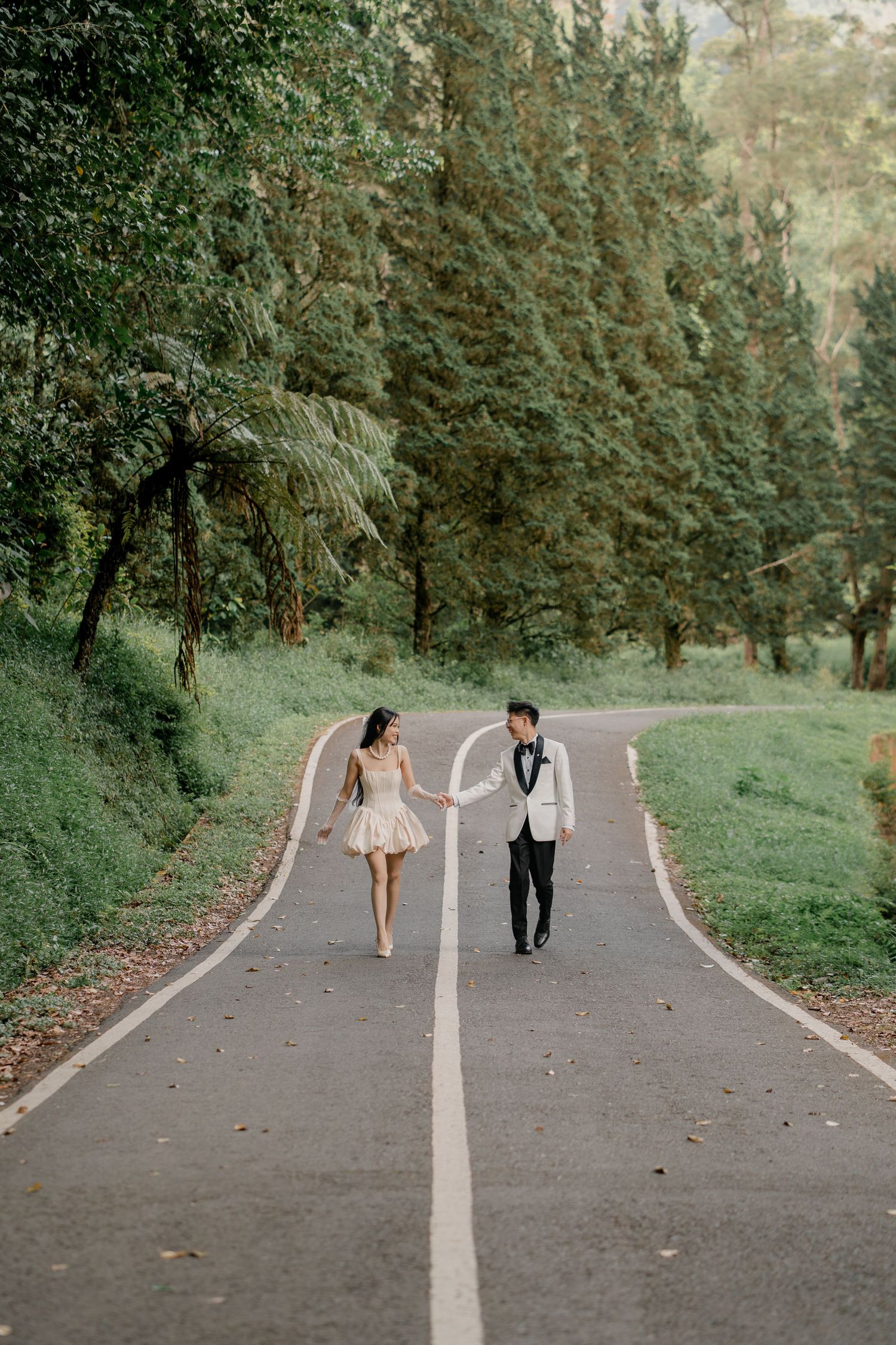 Couple holding hands walking on a winding road lined with lush green trees in formal wedding attire.
