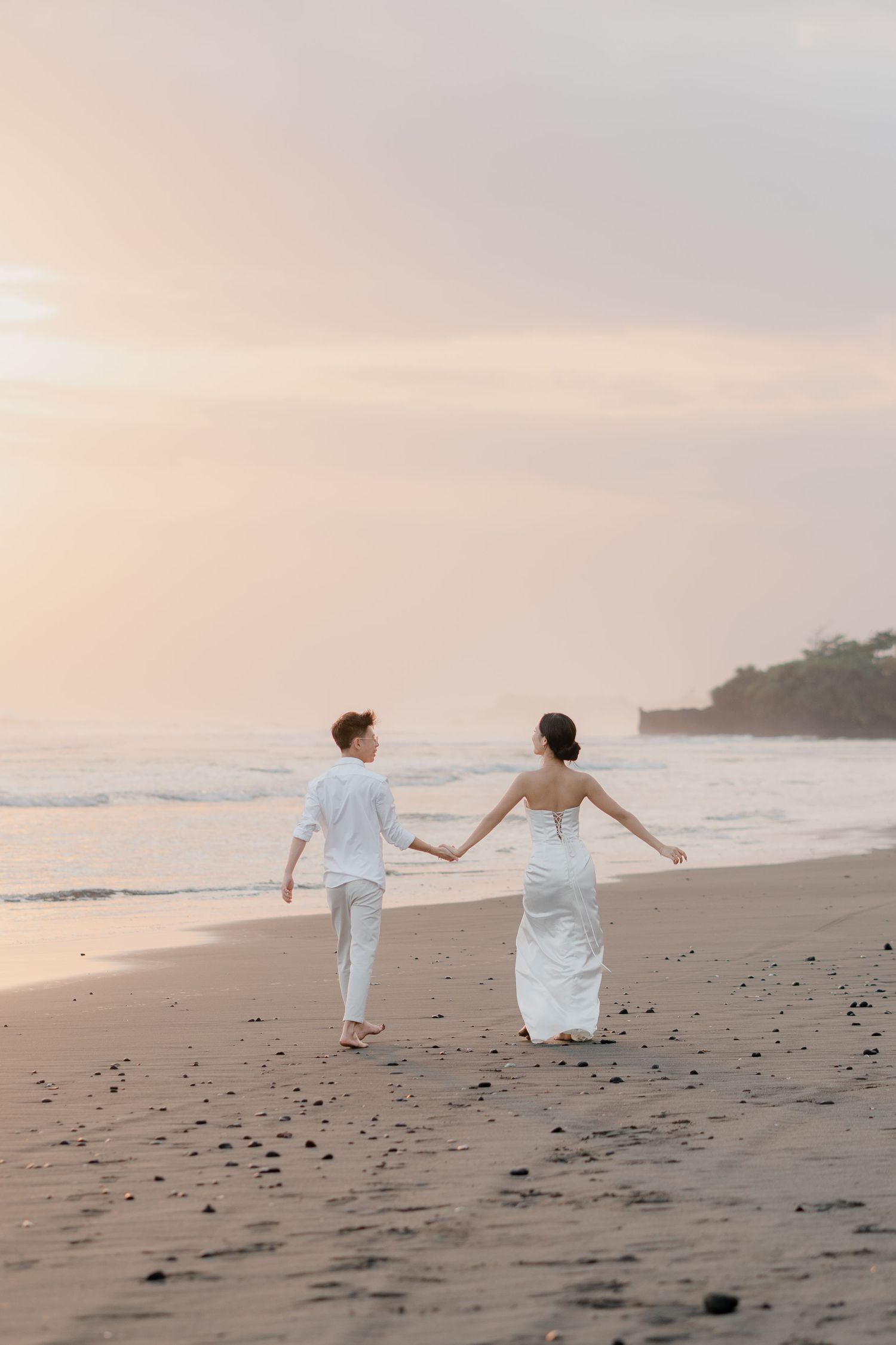 Couple in white outfits holding hands walking on a sandy beach at sunset with gentle ocean waves.
