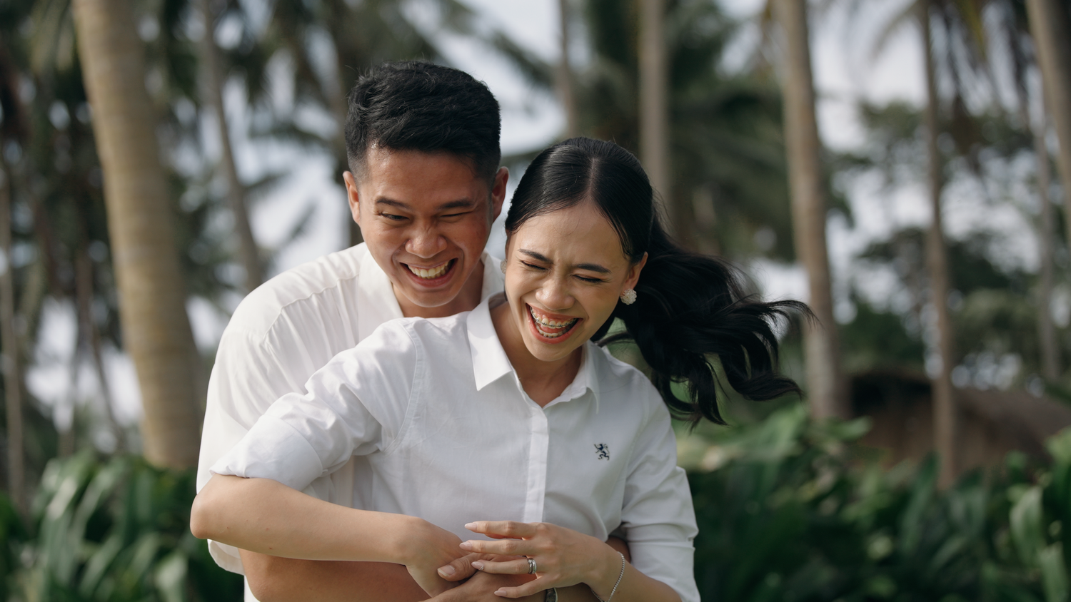 A smiling couple in white shirts embraces outdoors with tropical palm trees in the background.
