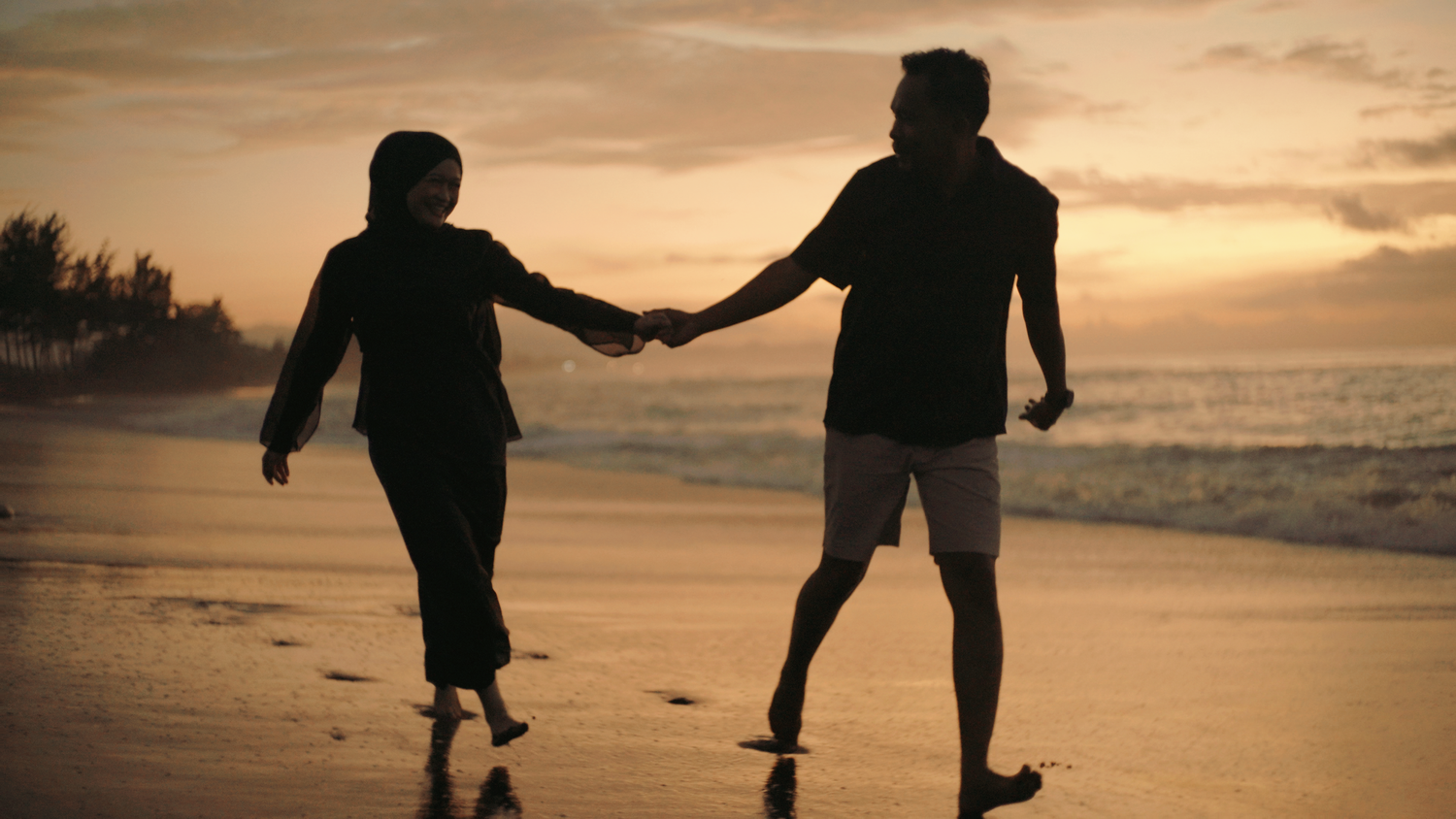 Silhouette of a couple holding hands and walking on a beach at sunset with golden sky and ocean waves.