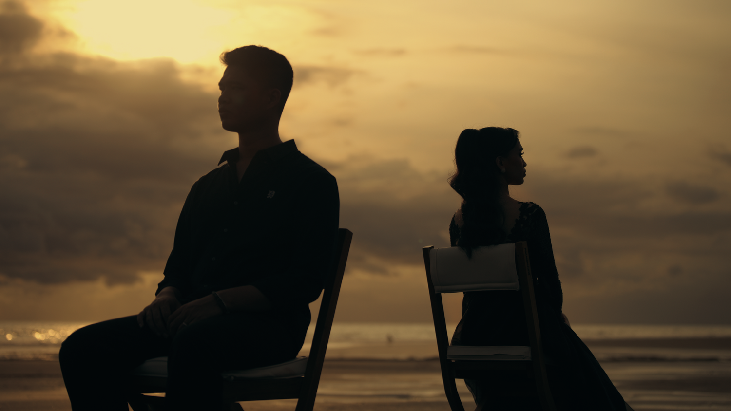 Two silhouettes sit back-to-back on chairs at sunset by the ocean, evoking themes of separation and longing.