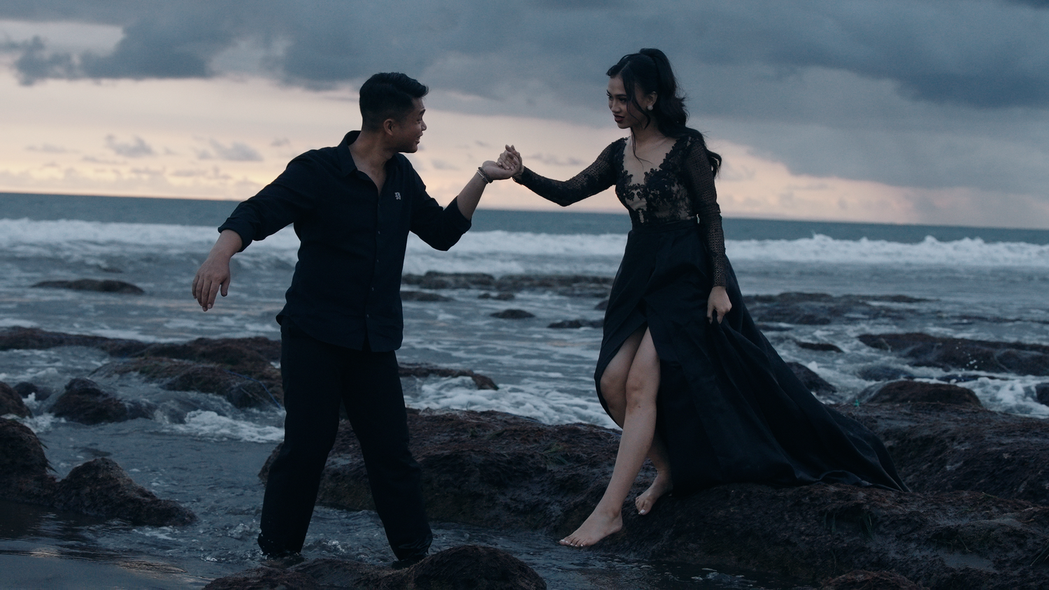 Couple dancing romantically on rocky beach at dusk, woman in flowing black gown, man in black outfit, ocean waves behind them.