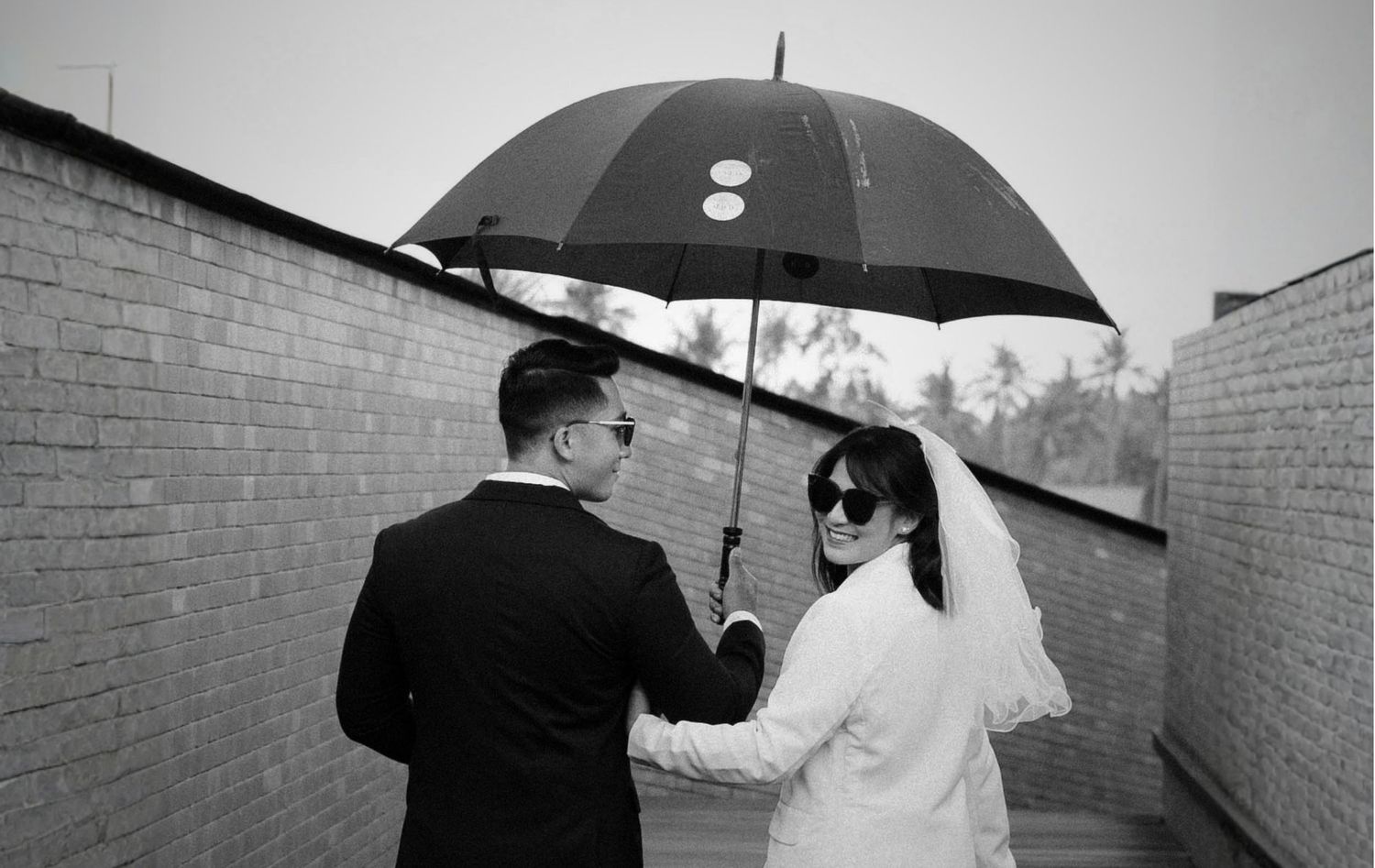 Black and white photo of a couple sharing an umbrella in the rain, bride in white dress and groom in suit.