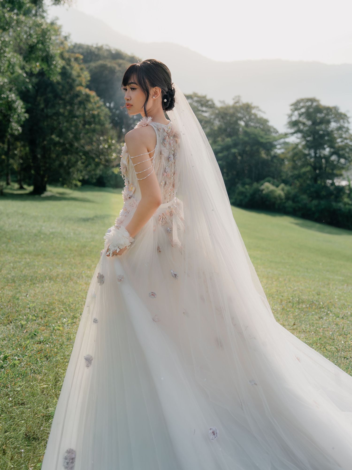Bride in elegant white tulle gown with floral details and long veil, standing on green lawn with mountain backdrop.
