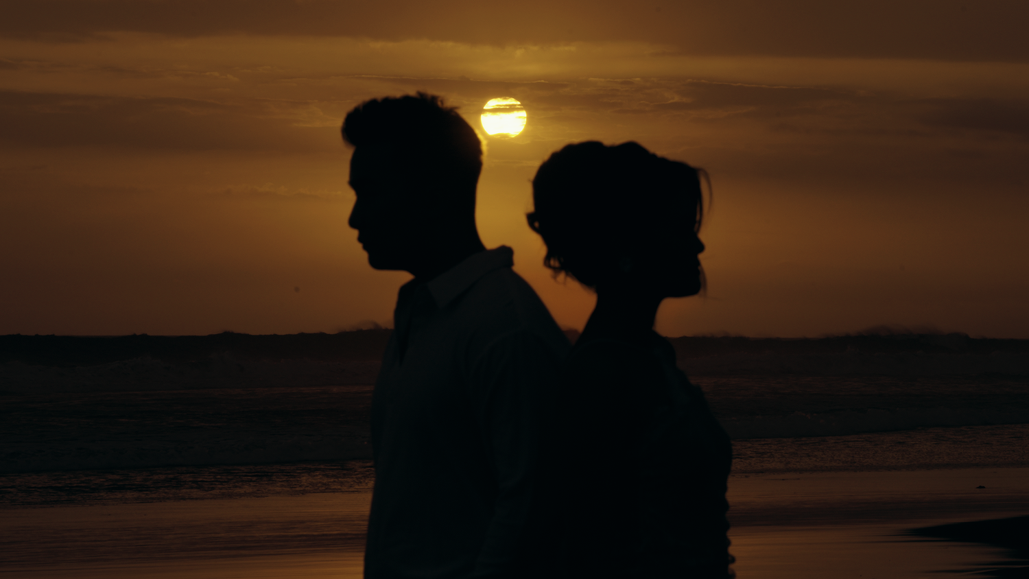 Silhouettes of a couple standing back to back on a beach at golden sunset with sun glowing on the horizon.