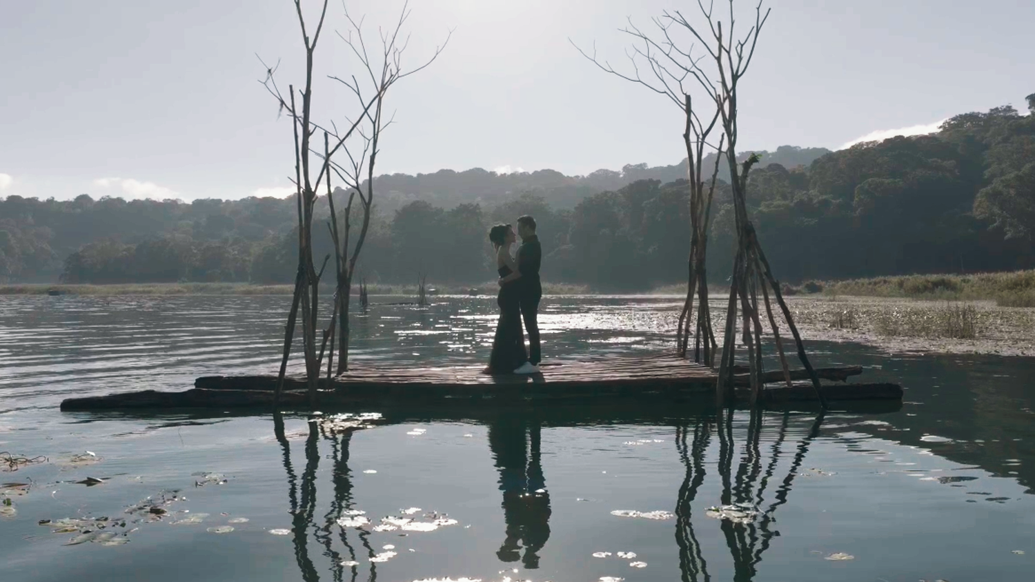 Couple standing on a small island in a reflective lake surrounded by bare trees and lush forest backdrop.