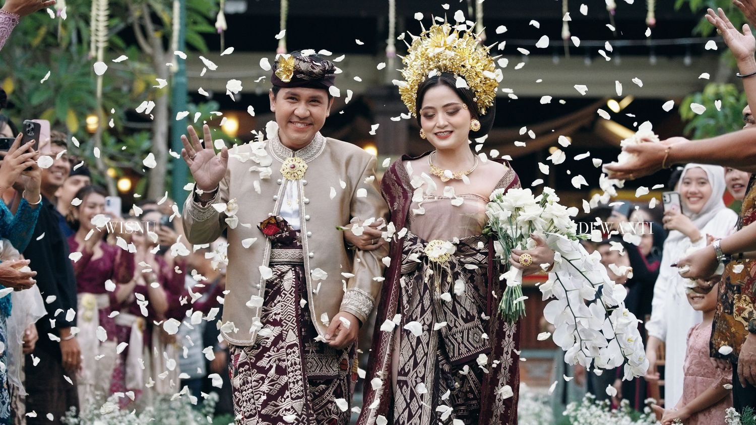 Bride and groom in ornate Javanese traditional wedding costumes celebrating with confetti.