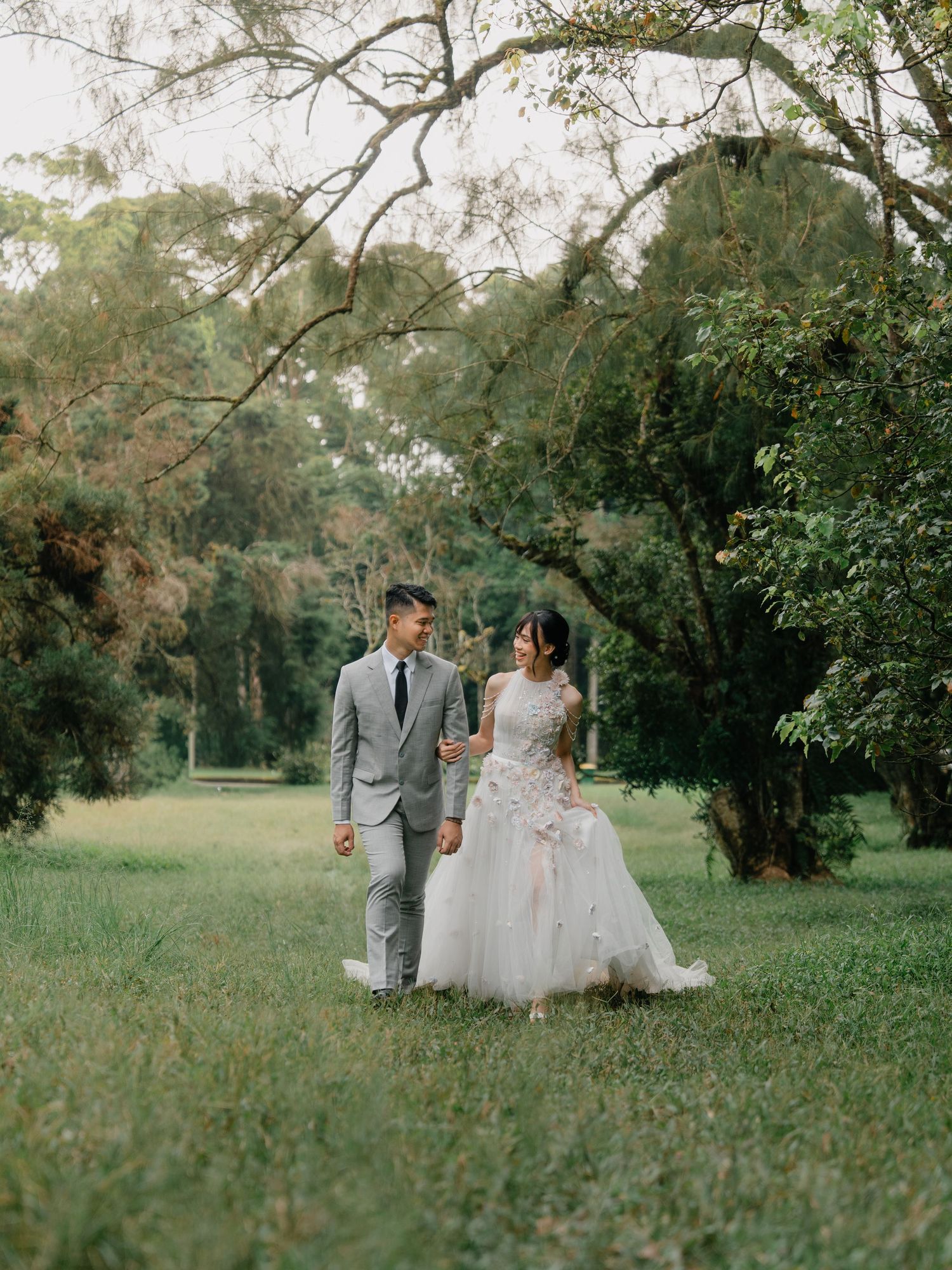 Bride and groom walking hand in hand through a lush green park, bride in floral gown, groom in gray suit.
