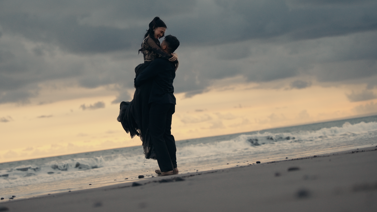 A couple embraces romantically on a beach at sunset, waves crashing behind them under a dramatic cloudy sky.