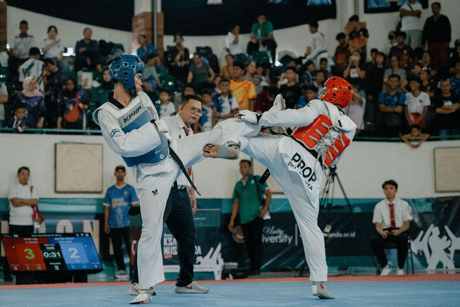 Two taekwondo athletes compete in a high-kick match wearing blue and red helmets at an indoor martial arts tournament.