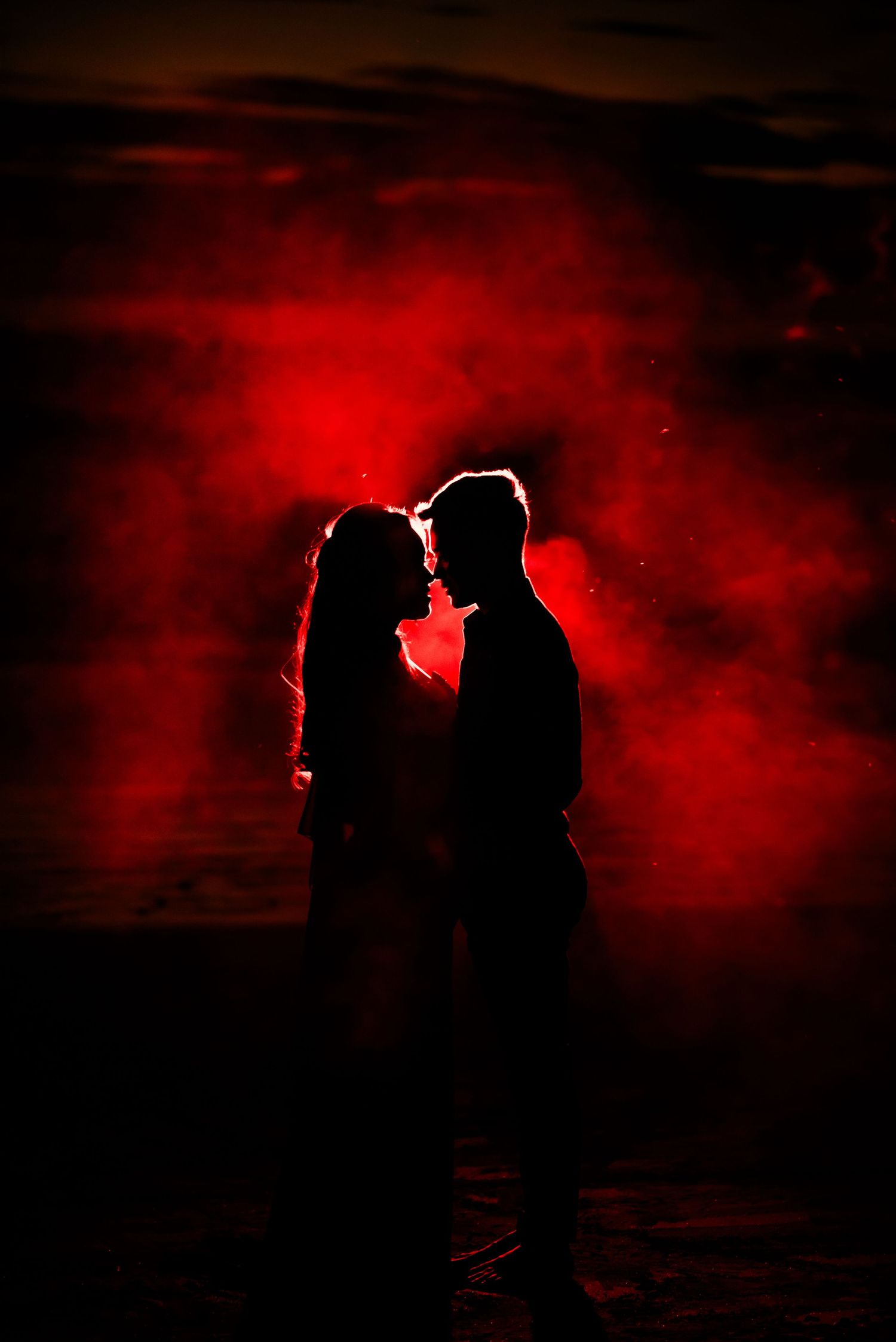 Romantic couple silhouette embracing against dramatic red smoke backdrop at night.