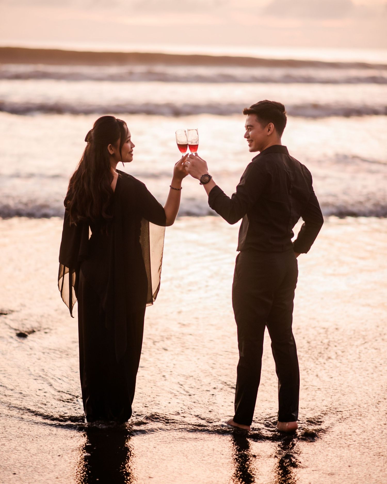 Couple in black attire toasting wine glasses on a beach at sunset with ocean waves in the background.
