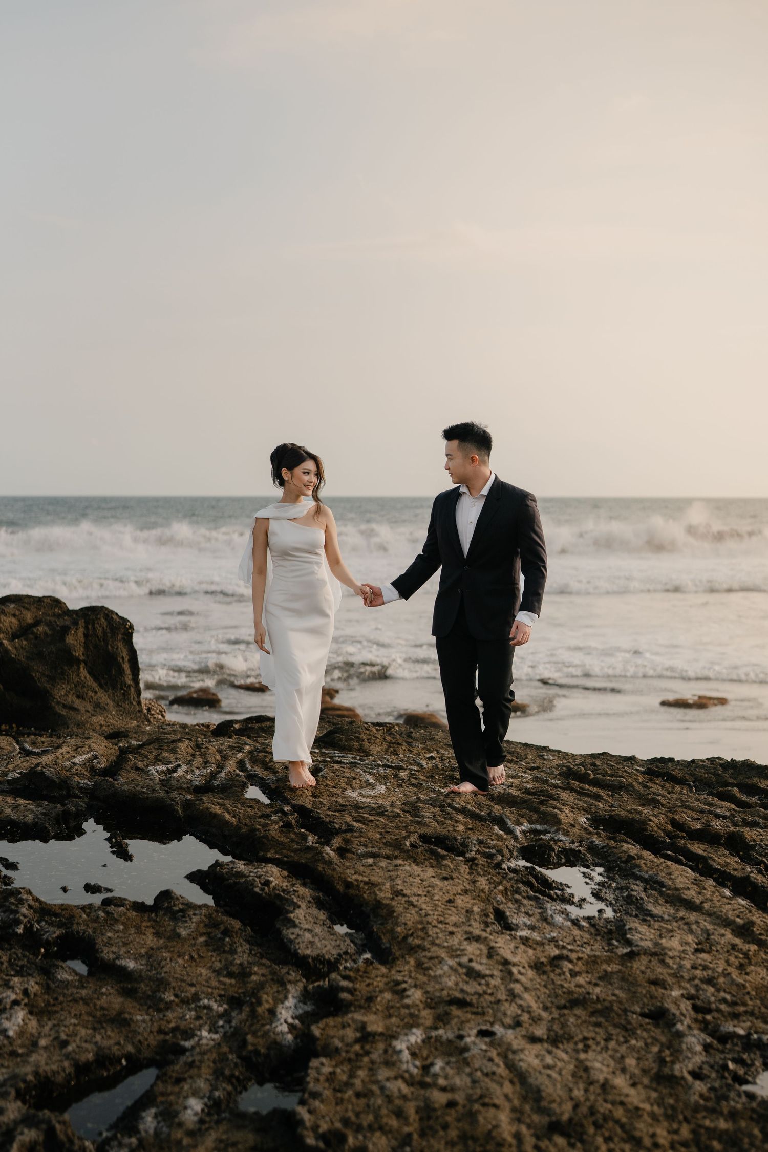 Couple in wedding attire holding hands on rocky beach with ocean waves, bride in white gown and groom in black suit.