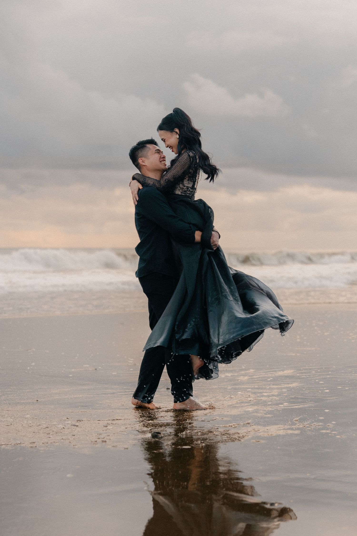 A couple in black attire embraces on a beach at sunset, man lifting woman as waves crash behind them.
