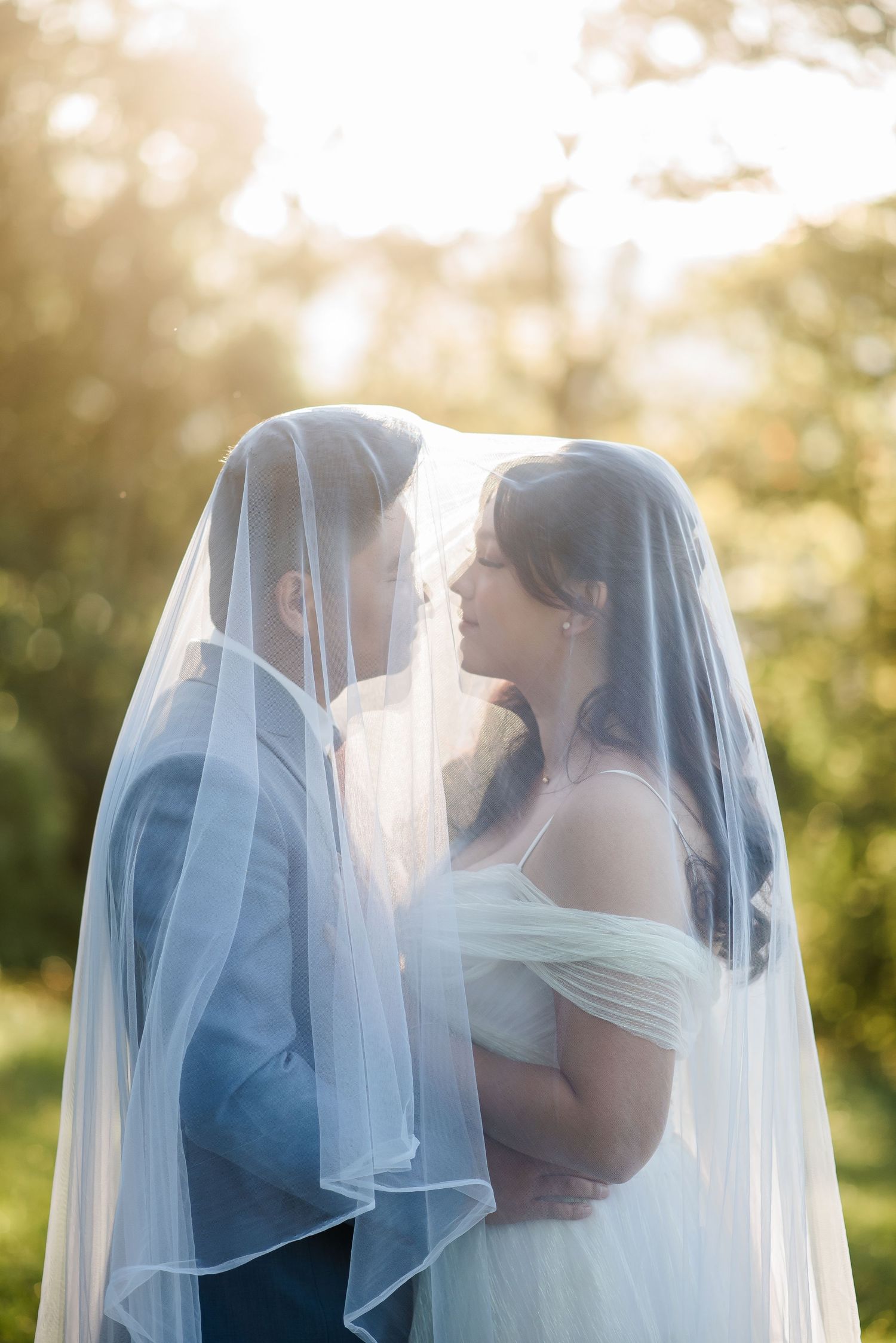 Bride and groom share an intimate moment under a sheer veil, bathed in golden sunlight outdoors.