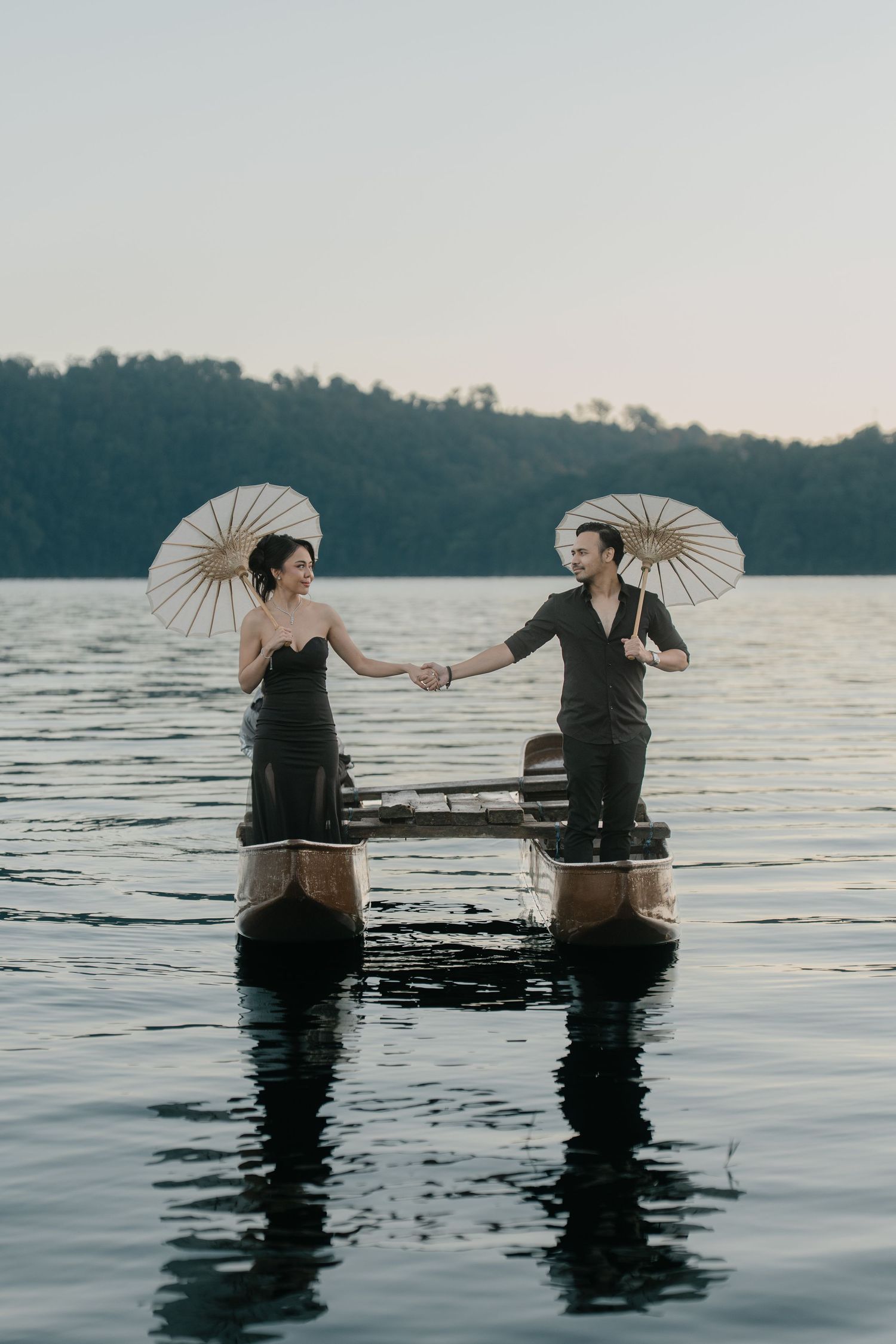 A couple holding hands on boats in a lake, each holding a white parasol, dressed in elegant black attire at dusk.