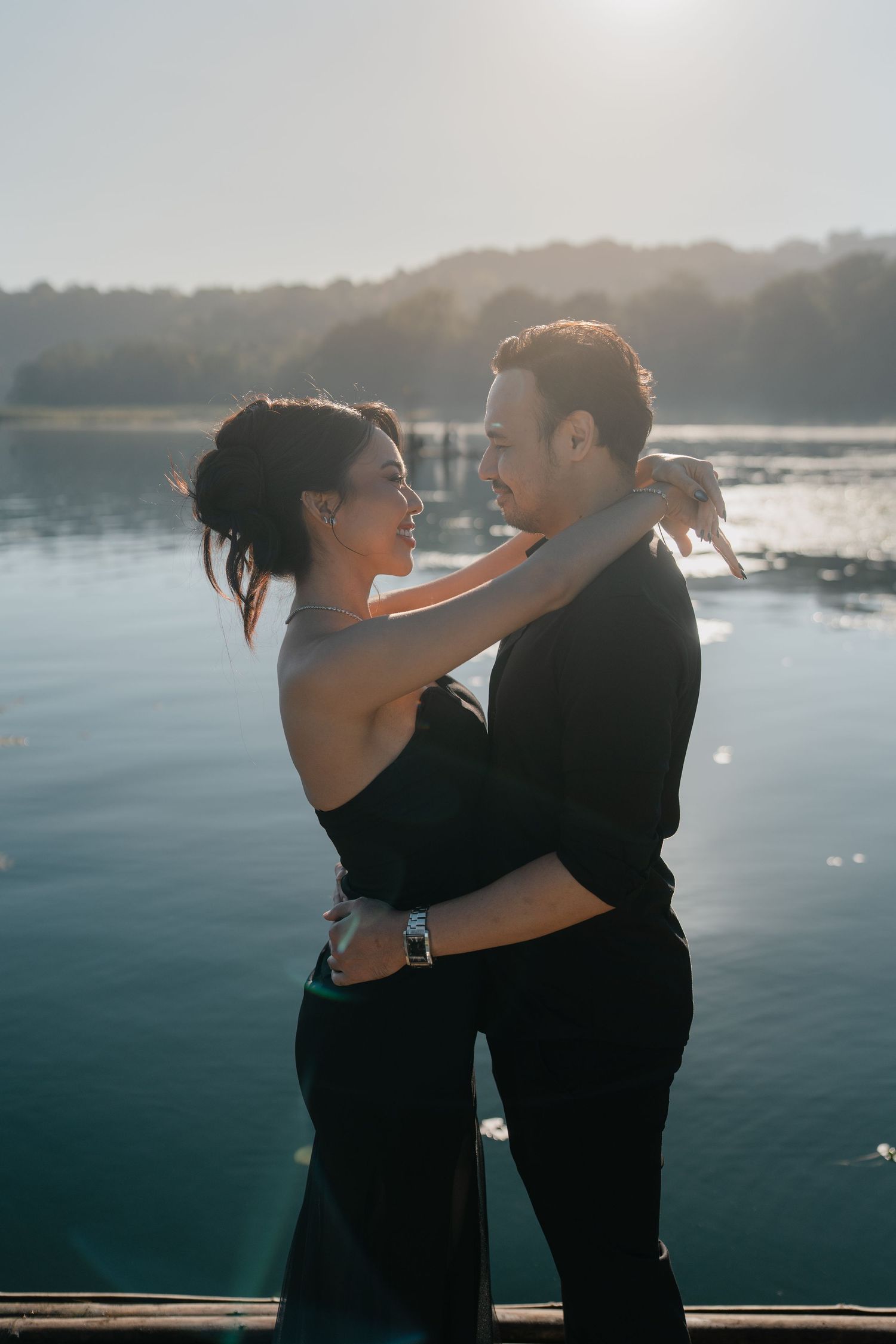 Couple embracing romantically by a sunlit lake, woman in black dress smiling at man in dark shirt outdoors.