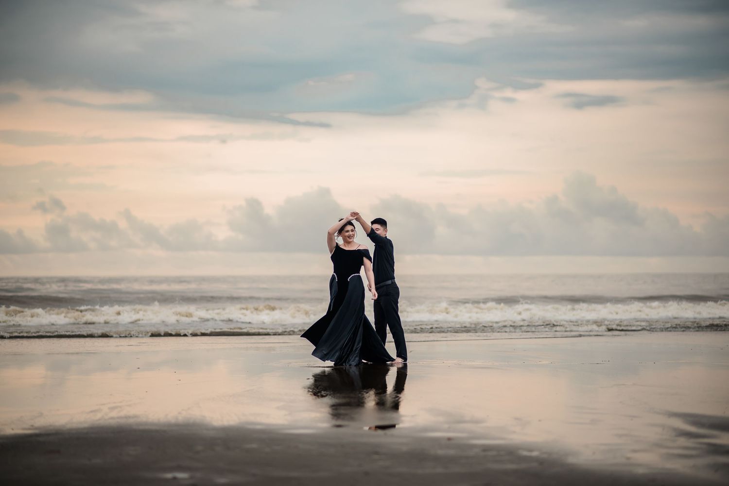 Couple dancing romantically on a reflective beach at sunset, wearing elegant black outfits with ocean waves in the background.