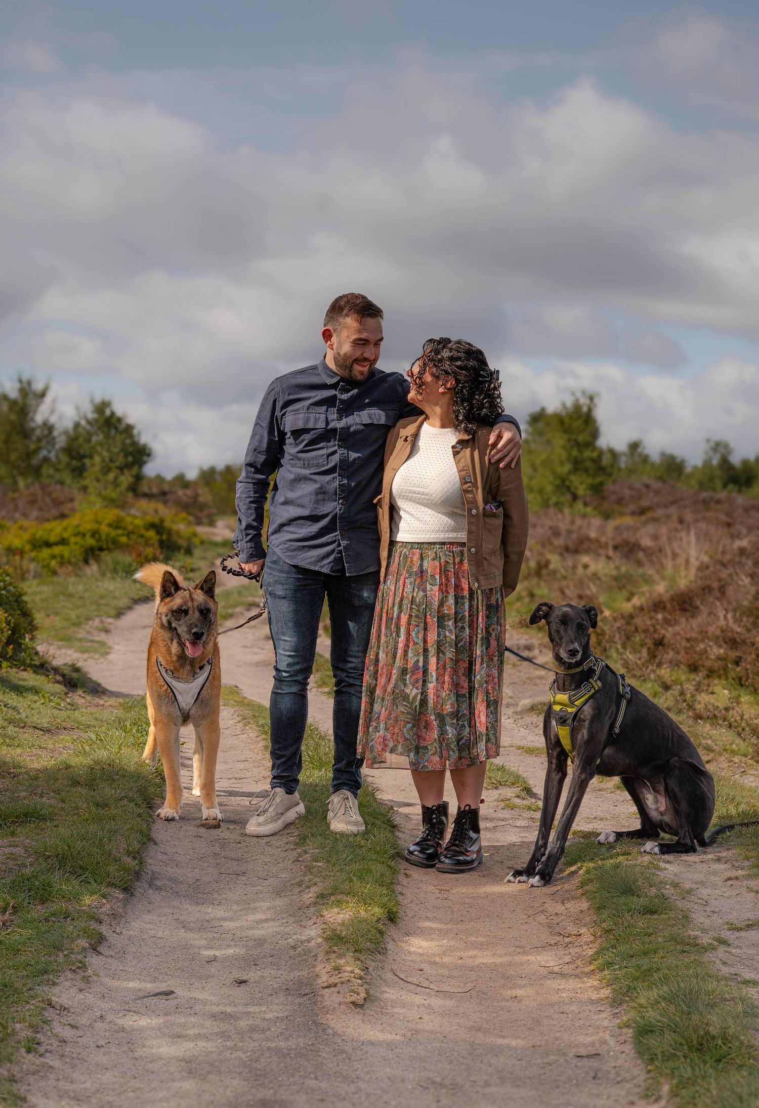 Couple walking two dogs on a scenic heathland path under a partly cloudy sky.
